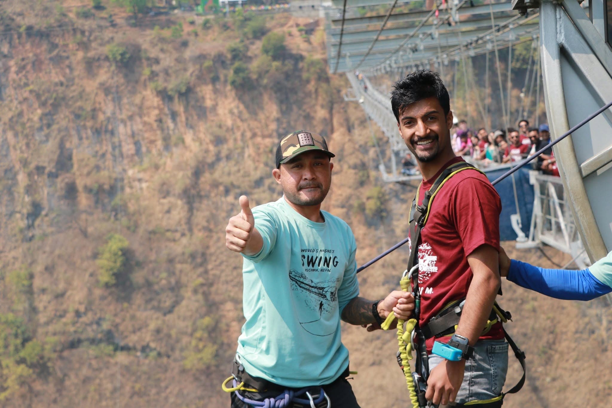 Subarna Poudel in a climbing harness at the Kushma swing, standing beside a staff member giving a thumbs up, with the gorge and suspension bridge behind them.