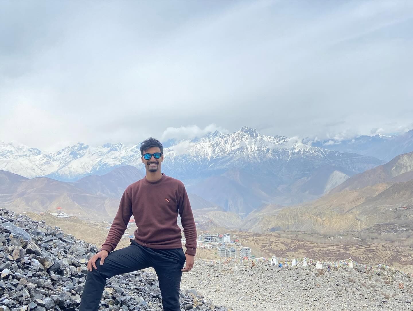 Subarna Poudel standing on a rocky ridge in front of a snow-covered Himalayan range.
