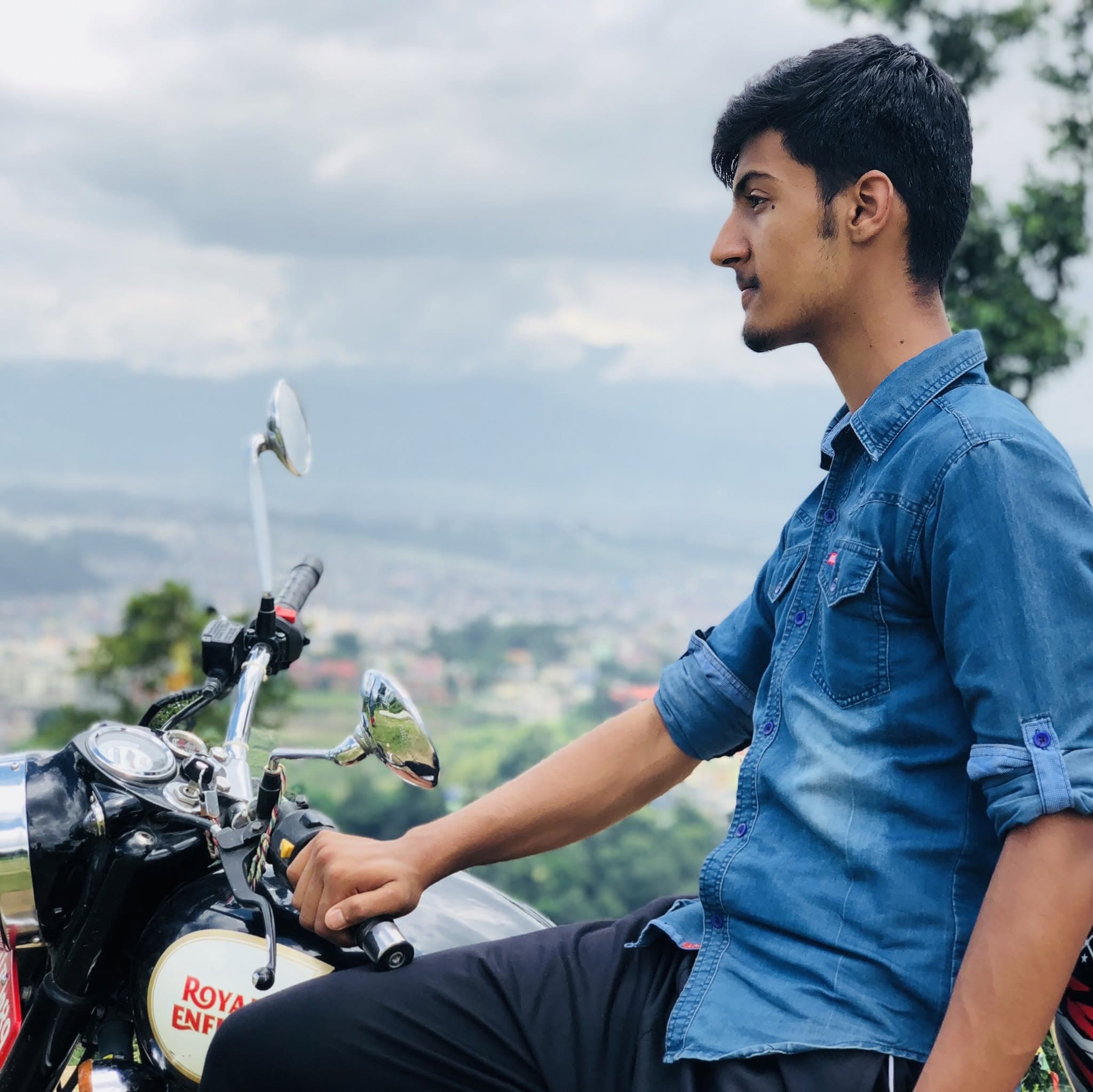 Subarna Poudel seated on a motorcycle at a hilltop viewpoint, looking out over a valley.