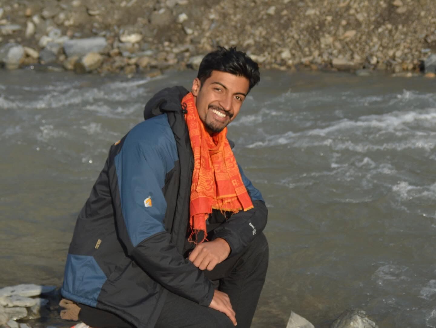 Subarna Poudel crouched on the stones beside a fast glacial river, wearing a winter jacket and an orange printed scarf.